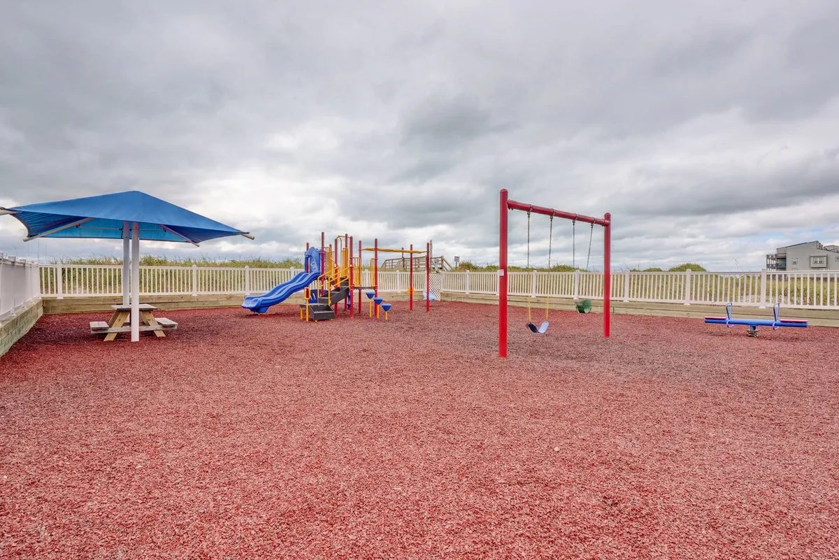 A cheerful playground scene featuring swings, slides, and a picnic table, set amidst bright colors and open space. Children are actively playing, and the area is designed for families and social gatherings.