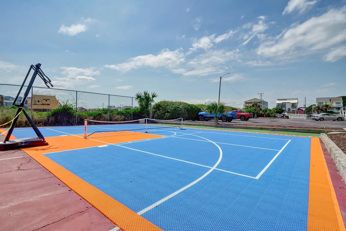 An exterior view of a multi-purpose sports court with a basketball hoop and a tennis net on a colorful court surface. The area is designed for recreational activities, such as basketball, tennis, and pickleball, supporting an active and healthy lifestyle.