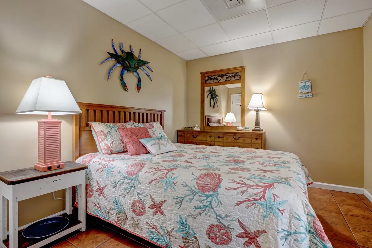 A cozy queen bedroom decorated with a coral-patterned quilt and playful crab wall art above the bed. Matching lamps on nightstands provide inviting, soft lighting, and the overall decor evokes a coastal, beach-inspired ambiance.