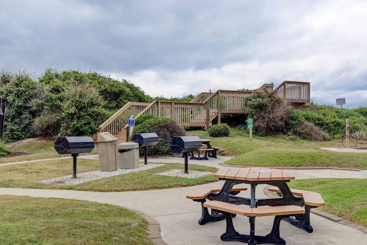 An outdoor area featuring barbecue grills and picnic tables, positioned near a staircase and surrounded by greenery. The setting is ideal for shared meals, socializing, and family gatherings after a day of play.
