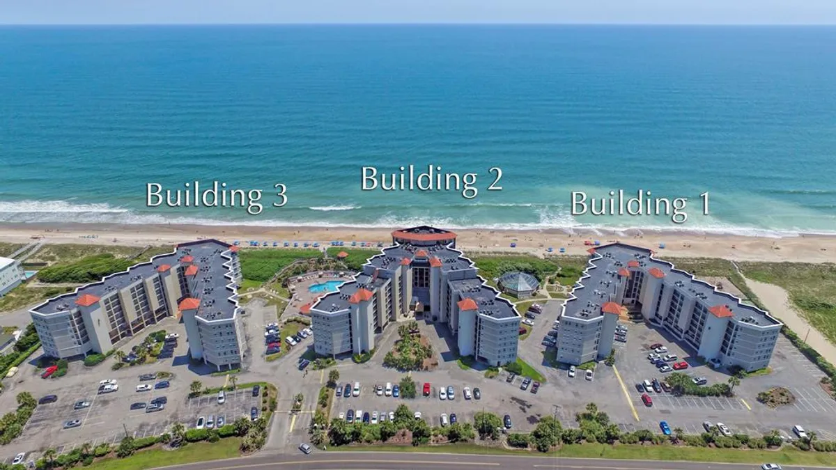 This exterior image captures three beachside resort buildings, a sparkling swimming pool, and the expansive ocean view beyond. The scene hints at a lively, social atmosphere with plenty of room for group activities, sunbathing, and easy access to both the beach and pool.