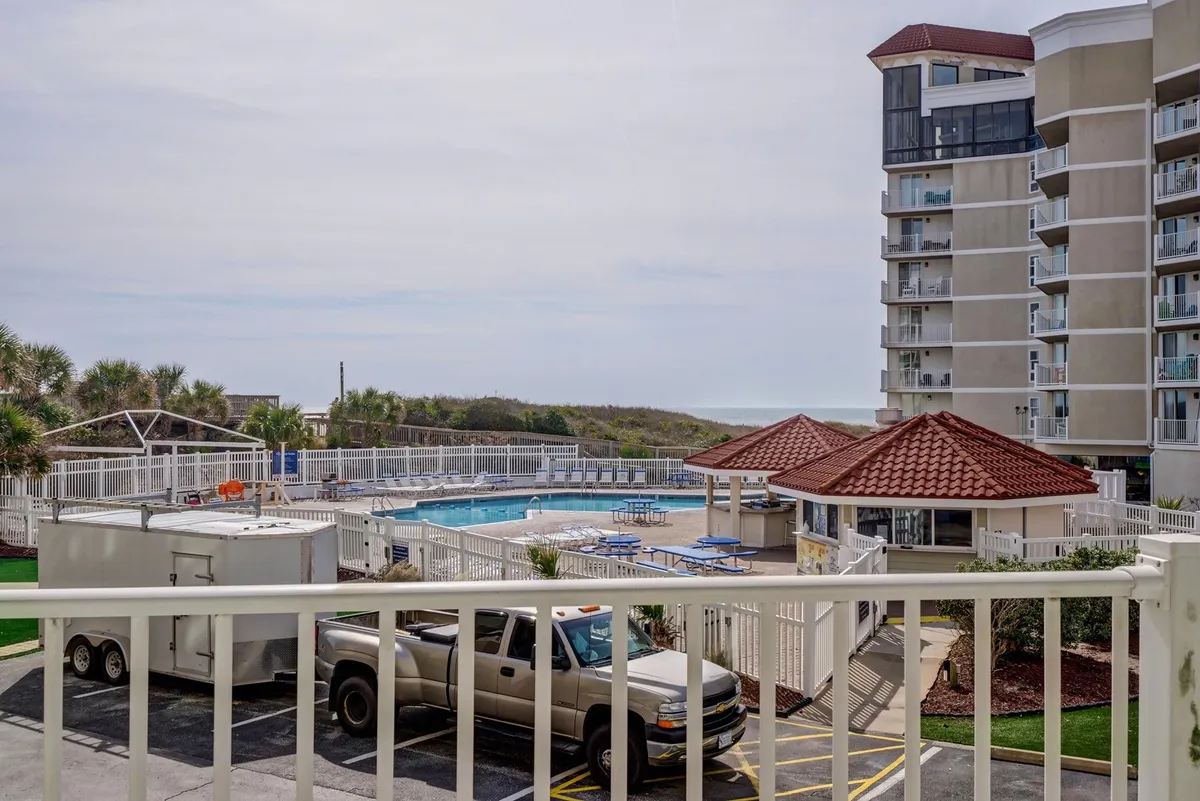 A vibrant exterior shot of a large swimming pool surrounded by palm trees, poolside seating, and resort-style building architecture. The scene evokes a sense of vacation and leisure, with ample sunlight and inviting spaces for swimming and relaxation.