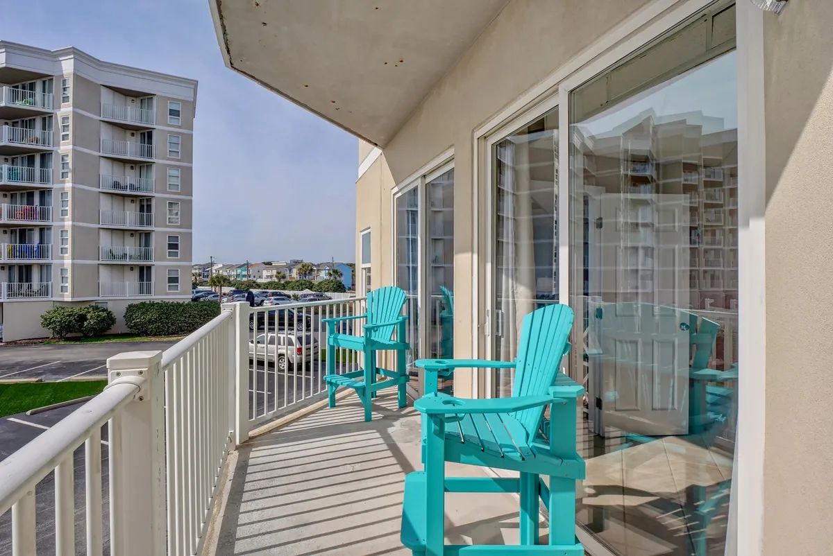 This image shows an inviting balcony with comfortable chairs positioned to enjoy the surrounding views. Glass doors connect the balcony to the interior, suggesting easy access and a seamless indoor-outdoor lifestyle. There is a clear view of the landscape, likely including glimpses of the ocean or pool area.