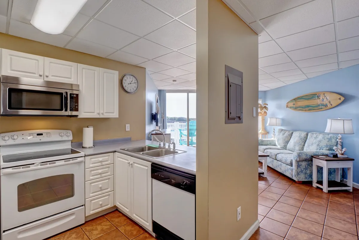 This image captures an open interior space combining a comfortable living area with a sofa, stylish surfboard decor, and a well-appointed kitchen. Ample sunlight streams through the windows, illuminating both the kitchen appliances and the casual seating area, evoking a breezy, coastal atmosphere ideal for socializing, cooking, and relaxation.