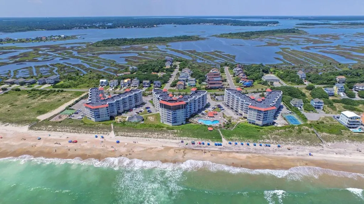 This image features the resort’s beachfront buildings, a sparkling swimming pool, the vast ocean waves beyond, and colorful beach umbrellas along the sand. Marshland and lush greenery frame the scene, creating an inviting coastal landscape.