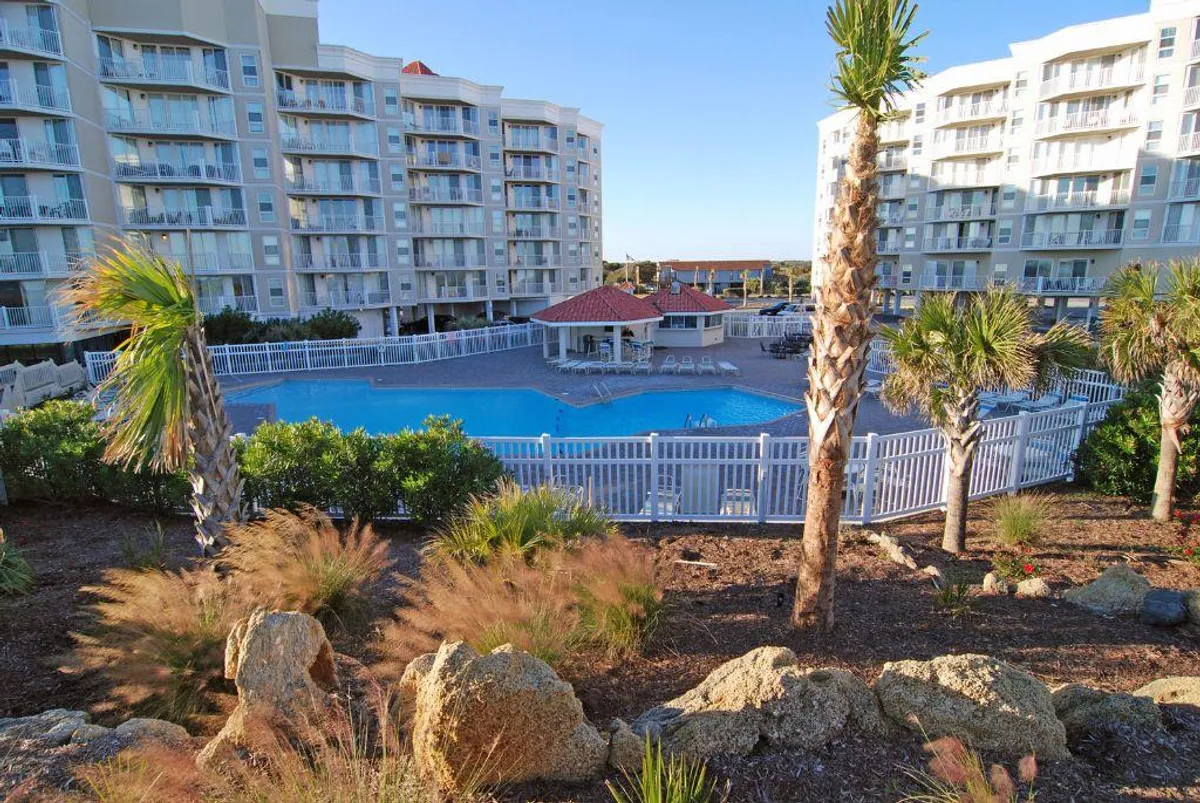 A vibrant exterior shot of the swimming pool surrounded by palm trees and lounge chairs, with residential buildings in the background. The pool area is lively and inviting, encouraging socializing, swimming, and relaxation under the sun.