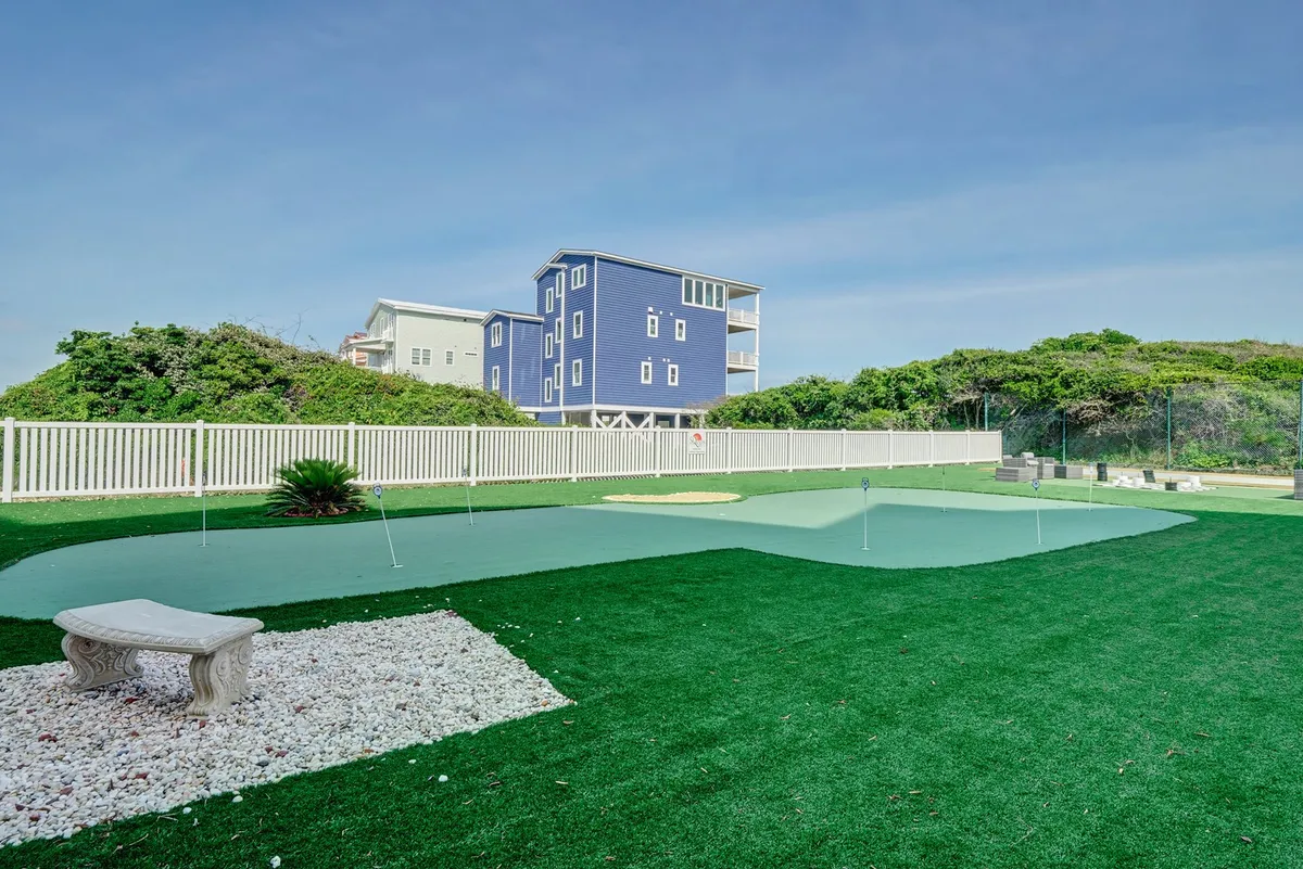 A vibrant exterior view of a green putting green bordered by a white stone bench and cheerful blue house. The scene invites social interactions, casual practice sessions, and moments of relaxation outdoors.