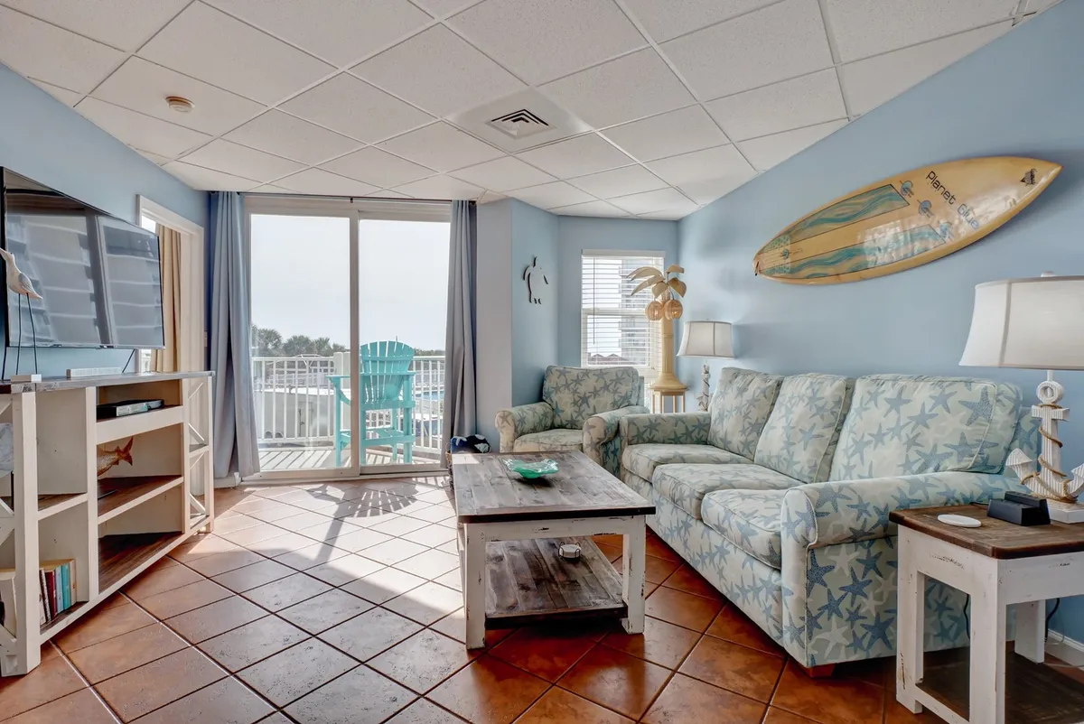 A cozy living area with colorful seating, large windows offering direct ocean views, and a surfboard decoration, creating a relaxed, beach-inspired vibe. The Smart TV is visible, making the space equally suited for entertainment and unwinding with friends or family.