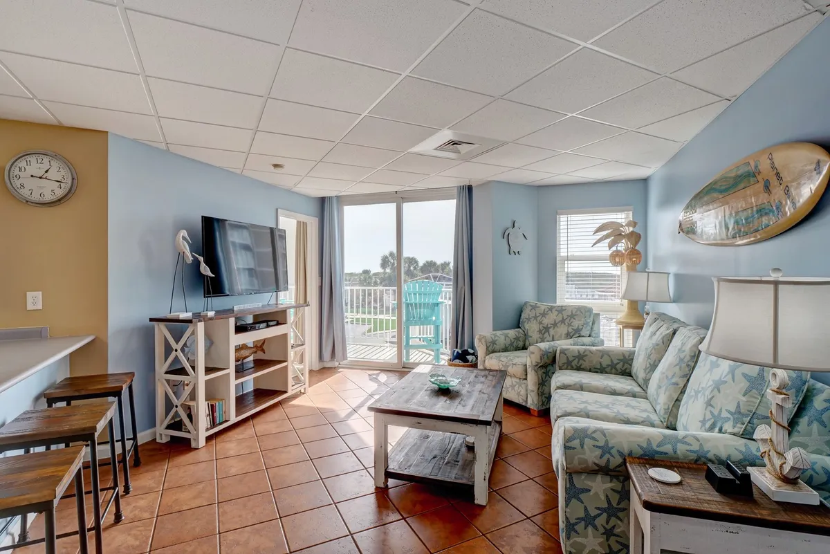 The image features a cozy living room with a starfish-patterned sofa, a coffee table, a TV, and a large window offering stunning sunrise views over North Topsail Beach. The inviting space is perfect for relaxing with family, enjoying movie nights, or savoring a morning coffee while taking in the coastal scenery.